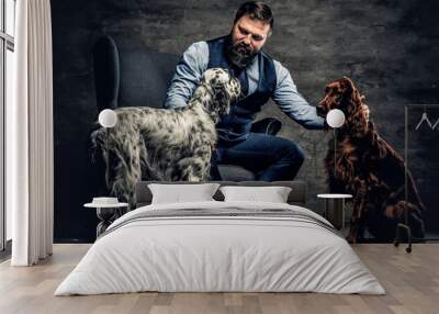 Middle-aged hunter dressed in elegant clothes sits on a sofa with his two purebred Setter dogs. Studio photo against a dark textured wall Wall mural