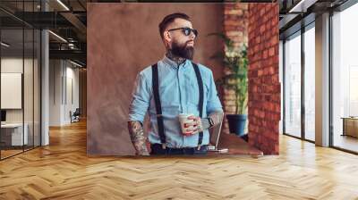 Old-fashioned tattooed hipster wearing a shirt and suspenders, in a sunglasses, standing with a cup of a coffee near a desk with a laptop, looking out the window in an office with a loft interior. Wall mural