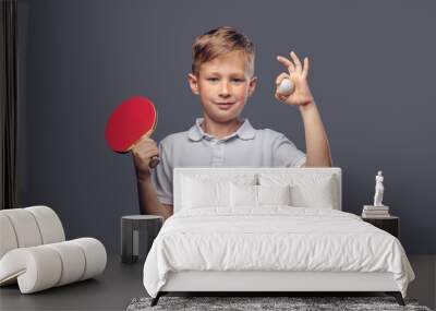 Redhead schoolboy dressed in a white t-shirt holds a ping-pong racquet and ball in a studio. Isolated on a gray background. Wall mural