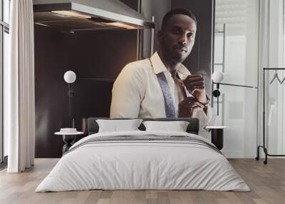 Sexy confident male in opened white shirt is putting on his wrist watch while sitting on the kitchen. Wall mural