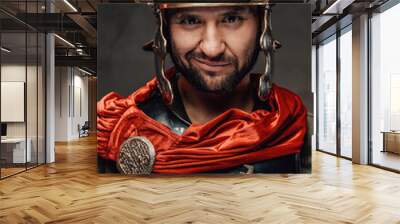 Smiley mature roman soldier in black armour with helmet and red mantle looking at camera in dark background. Wall mural