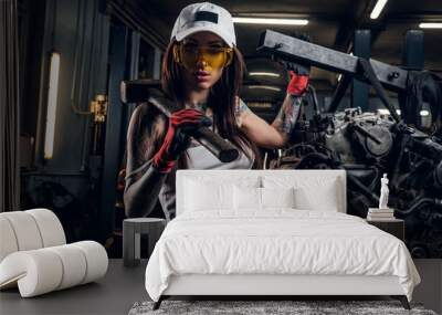 Stylish girl wearing cap and goggles posing with a steel hammer next to a car engine suspended on a hydraulic hoist in the workshop Wall mural