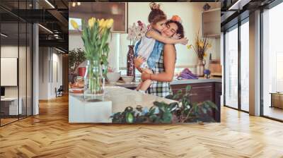 Sweet little girl kisses her mom while sitting in her arms in a kitchen. Wall mural