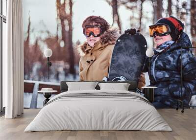 Two cheerful teenage brothers dressed in warm clothes sitting on a bench with a snowboard in a forest park Wall mural