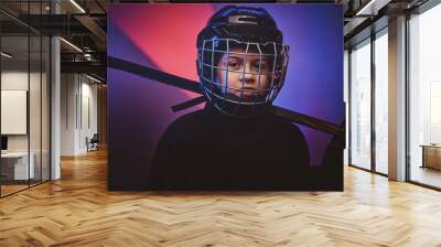 Young blonde sporty boy, ice hockey player, posing in a bright neon studio for a photoshoot, wearing an ice-skating uniform while wearing his helmet, holding a hockey stick and looking confident Wall mural