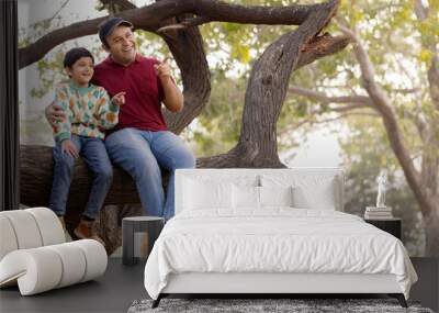 Cheerful father and son sitting on tree branch while admiring nature at park
 Wall mural