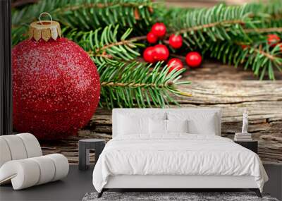 A red ornament sits on a wooden surface. The ornament is surrounded by green leaves and berries, giving the impression of a festive and joyful scene Wall mural