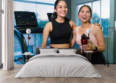 Two happy young fit indian friends women holding water bottle relaxing after workout at fitness club or gym, break time, healthy lifestyle, laughing, gossip, fun. Mental health. Stress free.  Wall mural