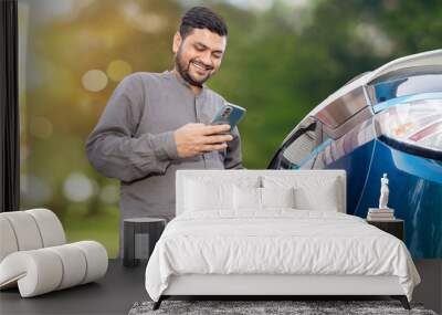 Happy Indian man attaching power cable into socket to charge his modern electric car at outdoor charging station while using smart phone. Electric vehicle Recharging battery charging port. Wall mural