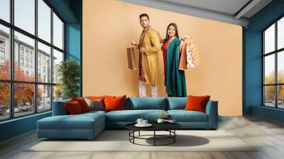 Happy young indian couple wearing traditional outfit holding shopping bags standing isolated on beige studio background. Diwali celebration and festive sale concept. Full length. Copy space. Wall mural