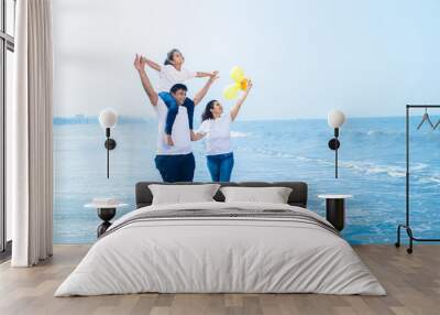 Happy young indian family of three wearing white t-shirt and blue jeans walking at beach, .Father carrying his daughter on shoulders, enjoying summer vacations. Travel and holidays. Copy space. Wall mural