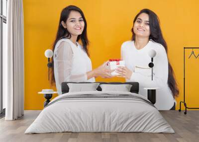 Happy young indian girls wearing white T-shirt exchange gift isolated over yellow orange background. studio shot. Festive celebration. Two happy brunette female friends exchanging presents, Wall mural