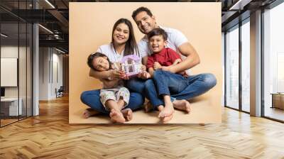 Happy young indian parents and kids wearing casual cloths holding house sitting together on floor isolated over beige studio background. Asian Family bonding. Concept of real estate and house buy. Wall mural