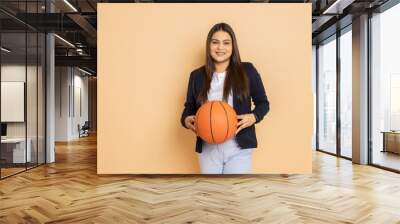 Portrait of beautiful young indian woman wearing formal suit holding basket ball in hand standing isolated over beige background. Wall mural