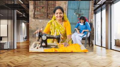 Portrait of happy traditional indian woman wearing sari using sewing machine while her young daughter studying behind her in school uniform. Wall mural