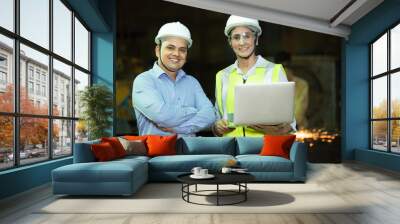 Portrait of two young happy Indian engineer male and female workers wearing safety white hard hat and vest holding laptop at industrial factory.Looking at camera with smile. Wall mural