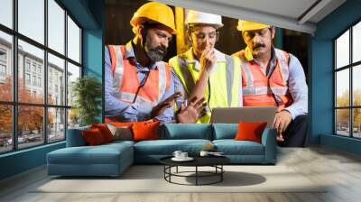 Team of young indian engineers wearing safety helmets and vest working together on a project at industrial site. Female engineer supervisor with laptop discussing plan and project, Wall mural