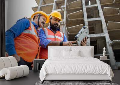 Two Indian male civil engineers or architect wearing helmet and vest holding digital tablet blueprint at construction site discussing real estate project. Low angle. Wall mural