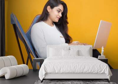 Young indian girl wearing white T-shirt using laptop while sitting on chair isolated over orange yellow background. Studio Shot, Copy space, Asian woman using computer.Technology concept. Wall mural