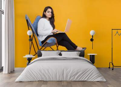 Young indian girl wearing white T-shirt using laptop while sitting on chair isolated over orange yellow background. Studio Shot, Copy space, Asian woman using computer.Technology concept. Wall mural