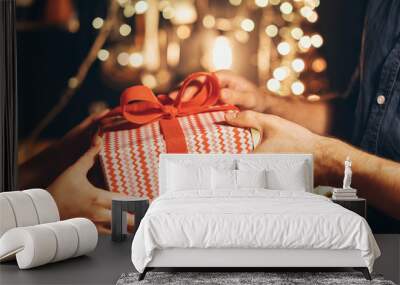 A woman gives a man a New Year's gift in red and white paper with a red bow. Hands of people with a New Year's gift. Lights in the background Wall mural