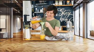 Attractive woman with a smile in her kitchen in a rustic style, makes fresh orange juice from a orange bottle by pouring it into a bottle. Housework. Vitamin drink. Wall mural