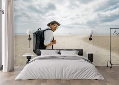 Traveler with a backpack in a white shirt with glasses and a hat barefoot walks along the sand on the beach. Wall mural