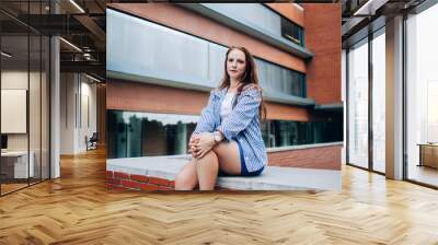 Portrait of charming redhead 20s woman in trendy outfit sitting next to urban setting building recreating on free time,attractive woman dressed in stylish apparel posing and looking at camera in town Wall mural