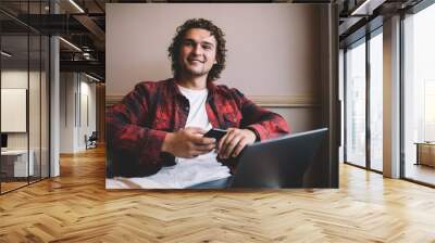 Smiling millennial man 20s connecting smartphone to laptop computer using bluetooth for share media files, positive male freelancer enjoying remote working process and modern gadgets for research Wall mural