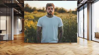 A young man with tattoos stands in a field at sunset, looking directly at the camera. Wall mural