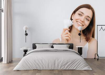 Beauty portrait of happy smiling woman with dark long hair with facial massager in hand on clean fresh skin face on the white background, banner Wall mural