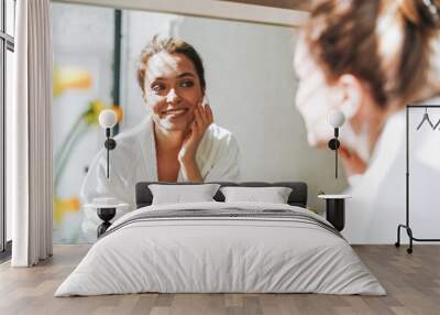 Beauty portrait of young smiling woman with dark long hair in white bathrobe put day nourishing moisturizer cream on clean fresh skin face and hands near mirror in bathroom at home Wall mural