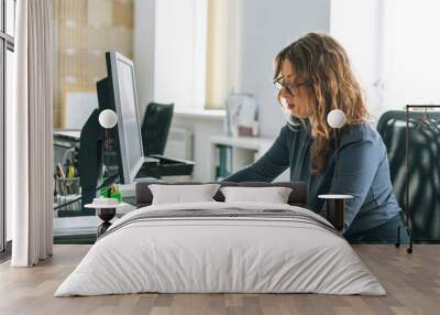 Charming smiling adult woman middle aged woman with curly hair architect designer working on computer in bright modern office Wall mural