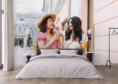 Happy smiling beautiful brunette young women friends in summer clothes with juice in hands walking on summer city street Wall mural