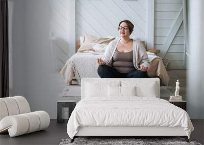 Middle aged plus size smiling brunette woman in home wear practicing yoga in bedroom Wall mural