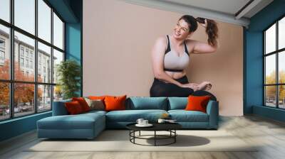 Self loving young woman plus size in sport wear doing stretching on beige background, body love Wall mural