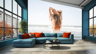 Young beautiful woman from back with long hair in beige sweater enjoying life on sea beach on sunset Wall mural