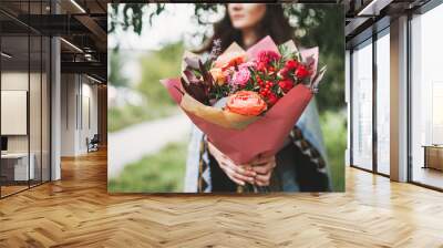 Young brunette woman in poncho and jeans holds a beautiful bouquet of different flowers in red tones Wall mural