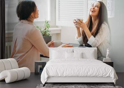 Young carefree brunette girls friends in casual with cups of tea meeting and sharing news in bright cafe or kitchen with green plants on table Wall mural