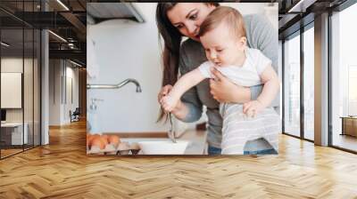 Young mother brunette woman with baby boy in arms cooking food in white modern kitchen at home Wall mural