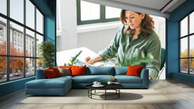 Young smiling brunette woman plus size working at laptop on table with house plant in the modern office Wall mural