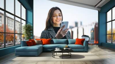 Young smiling woman in coat with coffee cup using mobile phone in evening city street Wall mural