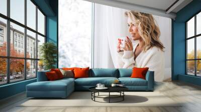 Young woman with blonde curly hair in white cardigan with cup of cocoa in hands looking at window with winter landscape at home Wall mural