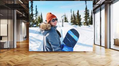 young charismatic girl snowboarder portrait in snowy forest in winter sunny day. Woman holding her snowboard and posing on top of the hill. Wall mural