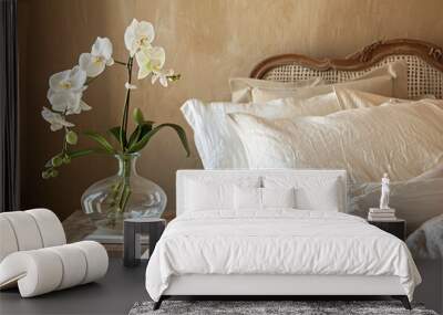 A bedroom with beige walls, featuring an antique wooden bed and white linen sheets on the headboard of which is placed a glass vase holding one orchid flower in full bloom. Wall mural