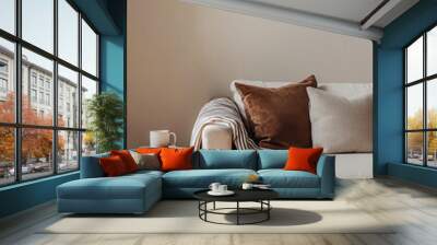 A beige sofa with pillows and a throw blanket on it in the living room, a side table with a coffee cup on the left side of the sofa, a carpet under the sofa, a beige wall behind the couch, minimalism. Wall mural