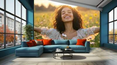 A happy curly-haired, dark-skinned young woman is smiling and enjoying nature with her arms outstretched outdoors in a park or meadow in summer. She is looking up at the sky while standing on a green  Wall mural