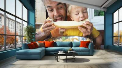 Family eating fresh sweet melon on terrace. Father and his toddler son biting together slice of honeymelon fruit. Summer food. Wall mural