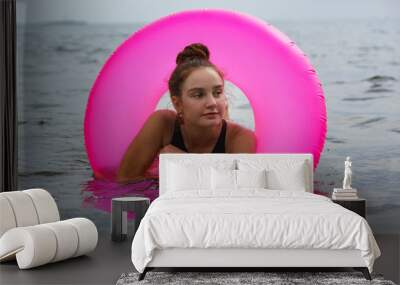 A beautiful teenage girl in a black swimsuit swims with a large inflatable pink swimming circle Wall mural