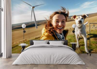 Smiling young attractive woman and her dog takes a selfie against the backdrop of a nature landscape with wind turbines. The concept of ecology, sustainable resources and climate optimism Wall mural